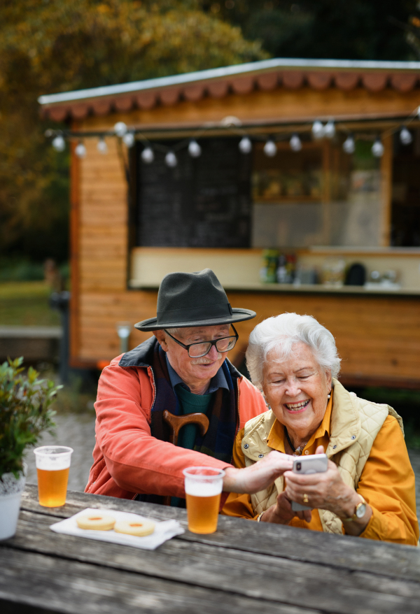 Happy senior couple in forest buffet resting after walk, having a beer and browsing on smartphone. Senior wife and husband looking at the photos on smartphone.