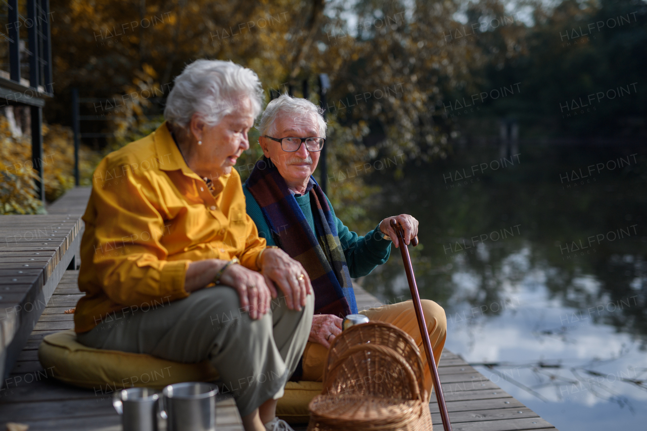 Happy senior couple in autumn clothes having break near a lake after walk.