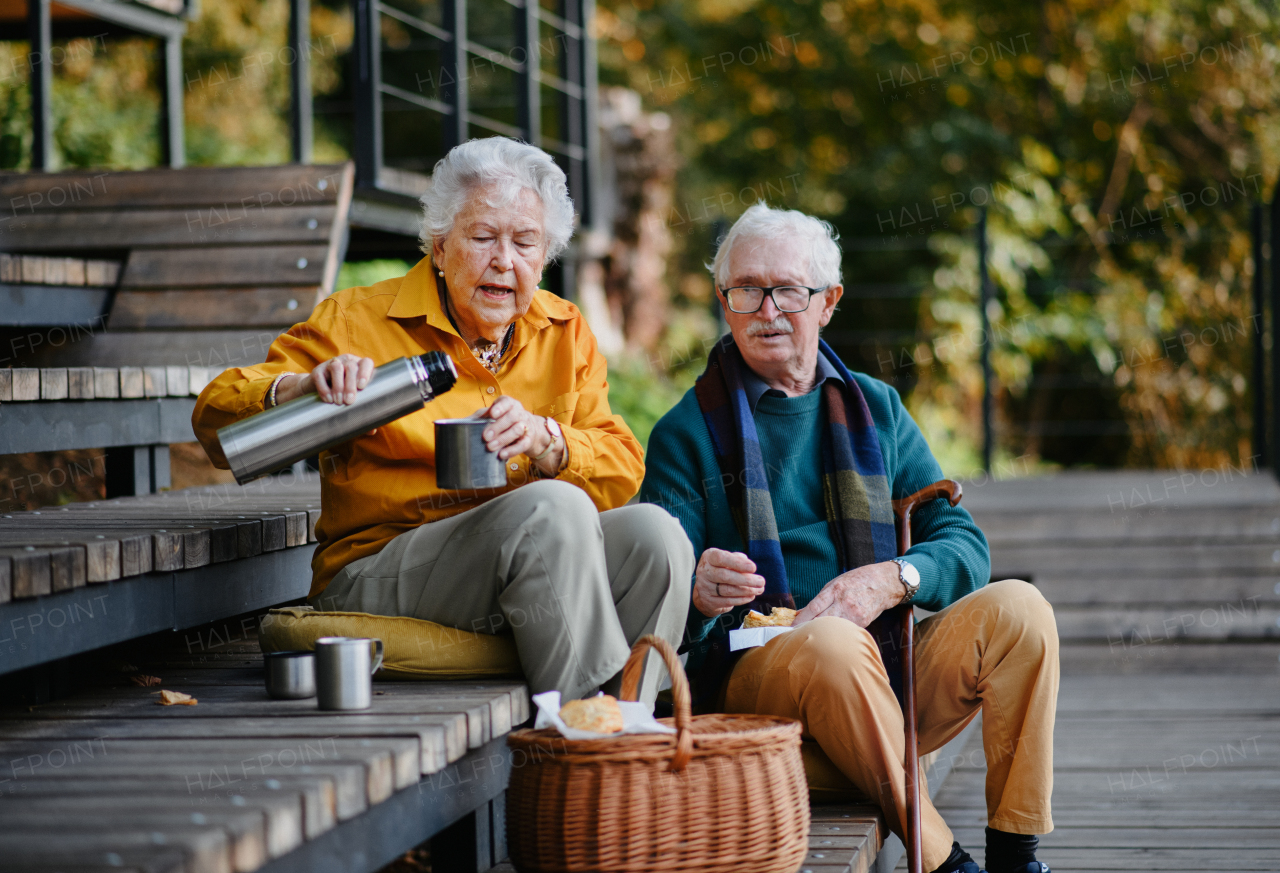 Happy senior couple having picnic and resting near lake after autumn walk. Wife serving tea from thermos bottle.