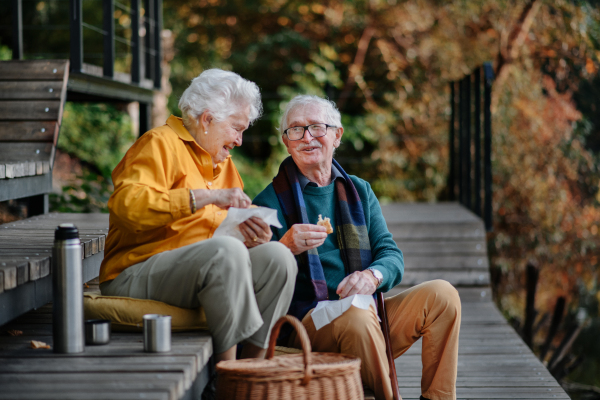 Happy senior couple in autumn clothes having picnic near a lake after walk.