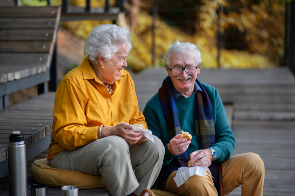 Happy senior couple in autumn clothes having picnic near a lake after walk.