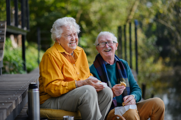 Happy senior couple having picnic and resting near lake after autumn walk.