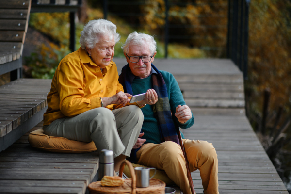 Happy senior couple resting near lake after walk and looking at a smartphone.