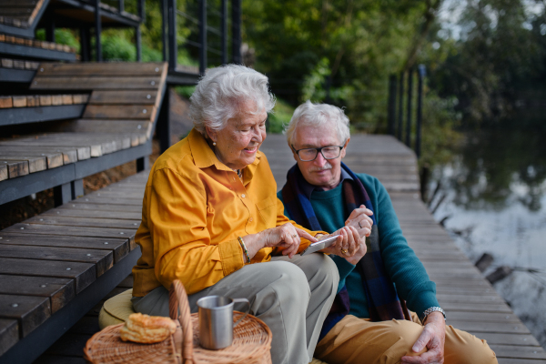 Happy senior couple resting near lake after walk and looking at a smartphone.