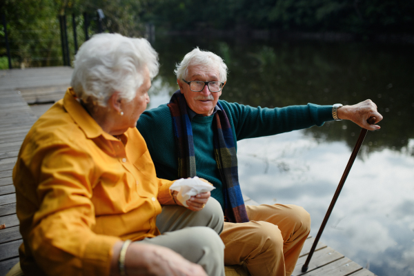 Happy senior couple in autumn clothes having break near a lake after walk.