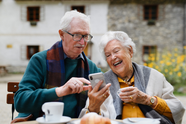 Happy senior couple in park cafe resting after walk, having a coffee and looking at smartphone.