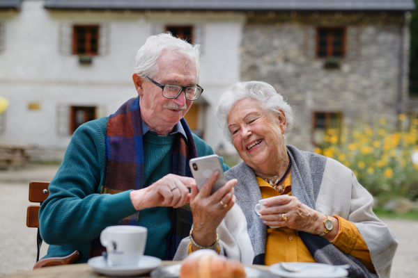 Happy senior couple in park cafe resting after walk, having a coffee and looking at smartphone.