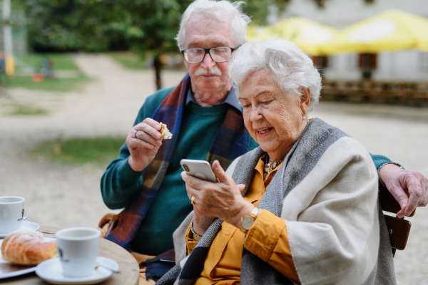 Happy senior couple in park cafe resting after walk, having a coffee and looking at smartphone.