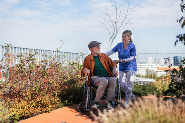 Nurse pushing senior man in a wheelchair. Female caregiver and elderly man enjoying a warm autumn day in nursing home, residential care home.