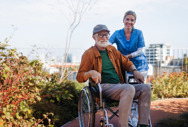 Nurse pushing senior man in a wheelchair. Female caregiver and elderly man enjoying a warm autumn day in nursing home, residential care home.