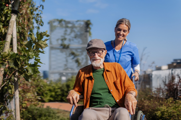 Nurse pushing senior man in a wheelchair. Female caregiver and elderly man enjoying a warm autumn day in nursing home, residential care home.