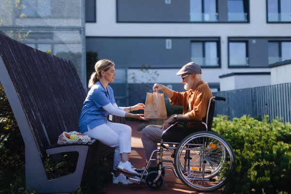 Nurse eating lunch or dinner outdoors with senior man in wheelchair. Female caregiver and elderly man enjoying a warm autumn day in nursing home, having picnic.