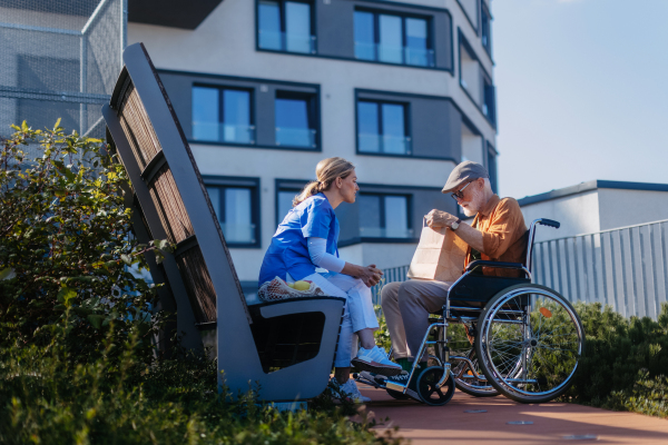 Nurse eating lunch or dinner outdoors with senior man in wheelchair. Female caregiver and elderly man enjoying a warm autumn day in nursing home, having picnic.