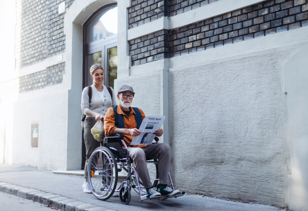 Granddaughter pushing senior man in wheelchair on street. Female caregiver and elderly man enjoying a warm autumn day, going home from shopping trip.