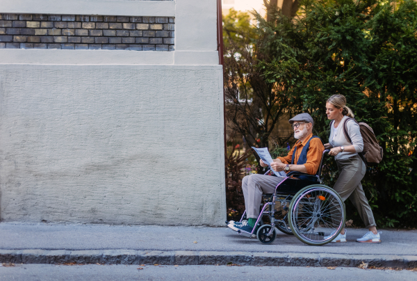 Granddaughter pushing senior man in wheelchair on street. Buying newspaper in newsstand. Female caregiver and elderly man enjoying a warm autumn day, going home from shopping trip.