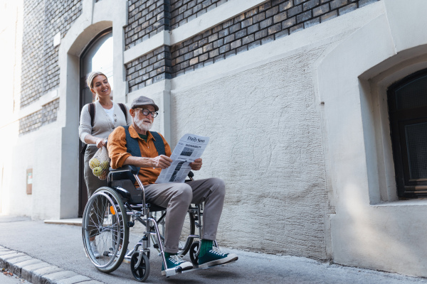 Granddaughter pushing senior man in wheelchair on street. Buying newspaper in newsstand. Female caregiver and elderly man enjoying a warm autumn day, going home from shopping trip.