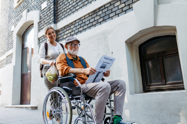 Granddaughter pushing senior man in wheelchair on street. Buying newspaper in newsstand. Female caregiver and elderly man enjoying a warm autumn day, going home from shopping trip.