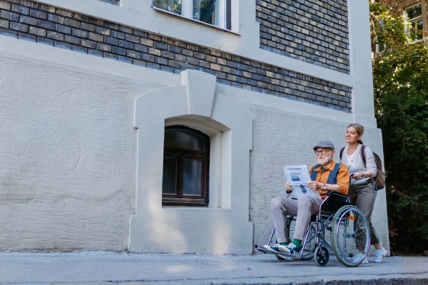 Granddaughter pushing senior man in wheelchair on street. Buying newspaper in newsstand. Female caregiver and elderly man enjoying a warm autumn day, going home from shopping trip.