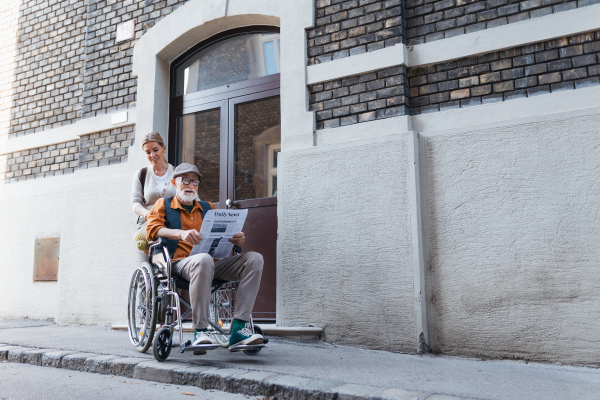 Granddaughter pushing senior man in wheelchair on street. Buying newspaper in newsstand. Female caregiver and elderly man enjoying a warm autumn day, going home from shopping trip.