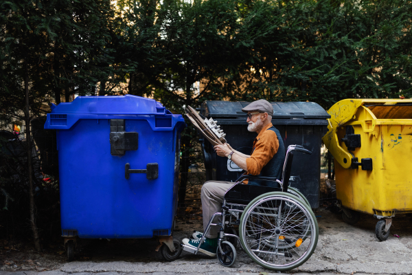 Senior man in wheelchair throwing paper waste, cardboard into recycling container in front his apartment complex. Elderly man sorting the waste according to material into colored bins.