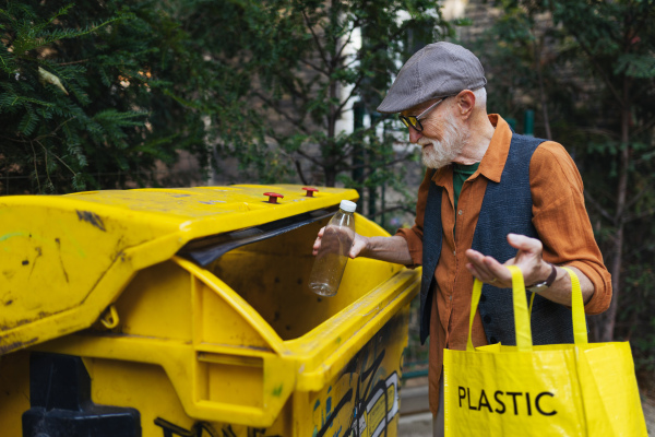 Senior man throwing plastic waste, bottles into recycling container in front his apartment. Elderly man sorting the waste according to material into colored bins.