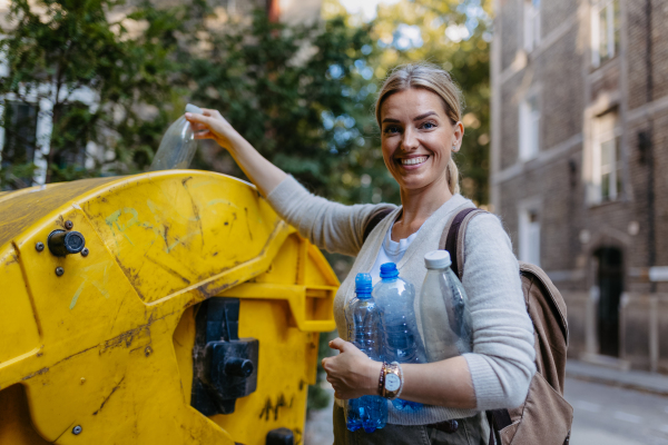 Woman throwing plastic waste, bottles into recycling container in front her apartment complex. Woman sorting the waste according to material into colored bins.