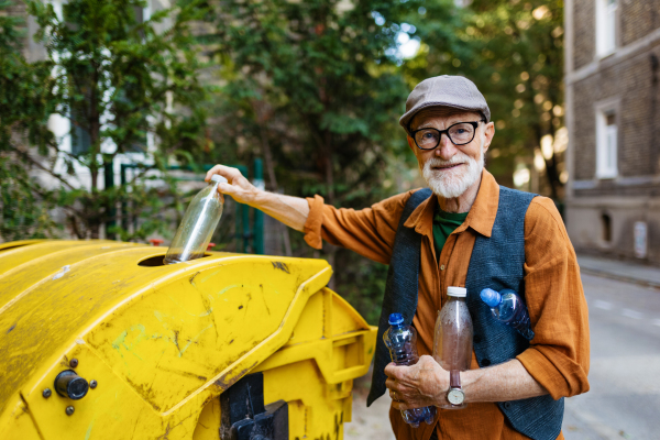 Senior man throwing plastic waste, bottles into recycling container in front his apartment. Elderly man sorting the waste according to material into colored bins.