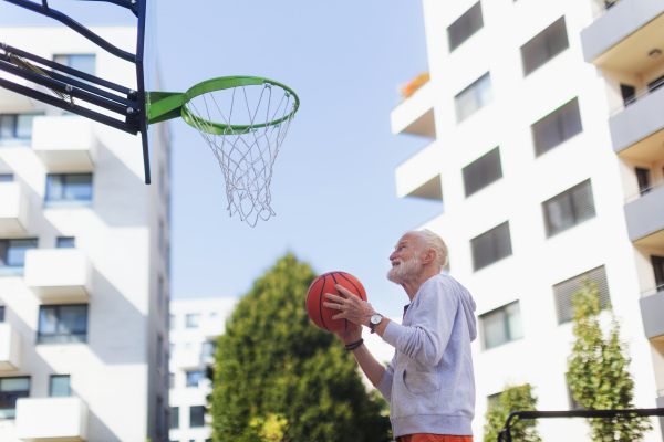 Senior man playing basketball outdoors on basketball court in the city, in community center. An older, vital man has active lifestyle, doing sport every day.