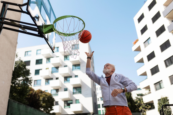 Senior man playing basketball outdoors on basketball court in the city, in community center. An older, vital man has active lifestyle, doing sport every day.