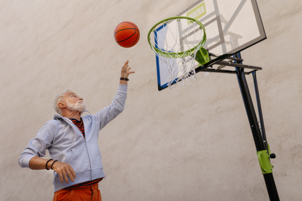 Senior man playing basketball outdoors on basketball court in the city, in community center. An older, vital man has active lifestyle, doing sport every day.