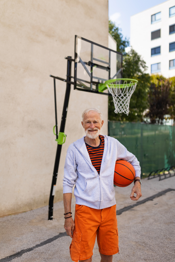 Senior man playing basketball outdoors on basketball court in the city, in community center. An older, vital man has active lifestyle, doing sport every day.