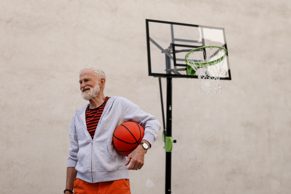 Senior man playing basketball outdoors on basketball court in the city, in community center. An older, vital man has active lifestyle, doing sport every day.