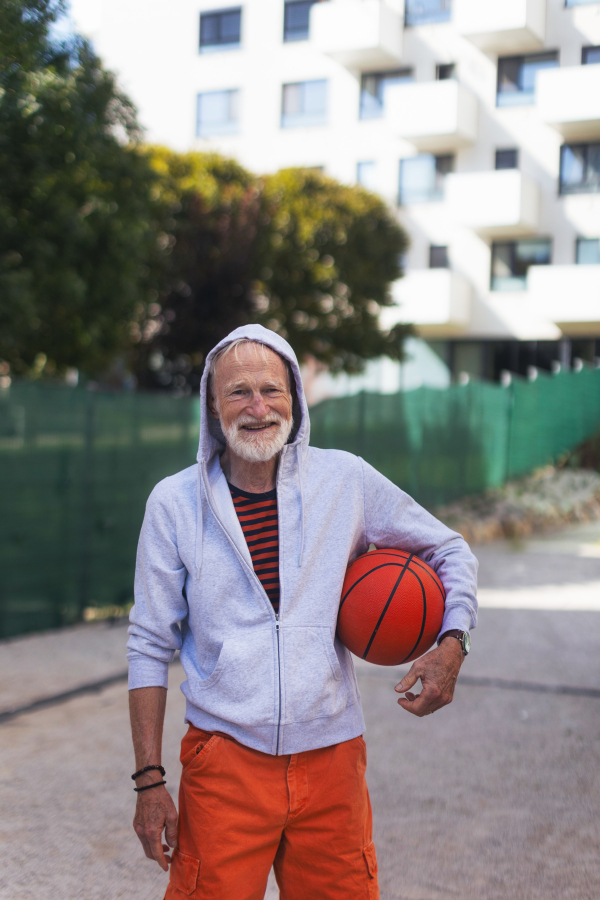 Senior man playing basketball outdoors on basketball court in the city, in community center. An older, vital man has active lifestyle, doing sport every day.