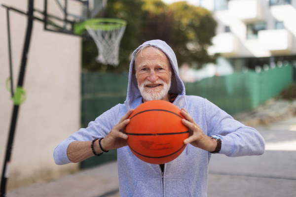 Senior man playing basketball outdoors on basketball court in the city, in community center. An older, vital man has active lifestyle, doing sport every day.