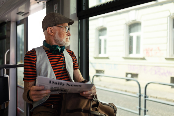 Elderly man traveling through the city by bus, reading a newspaper. Senior city commuter taking tram to grocery store, using public transportation.