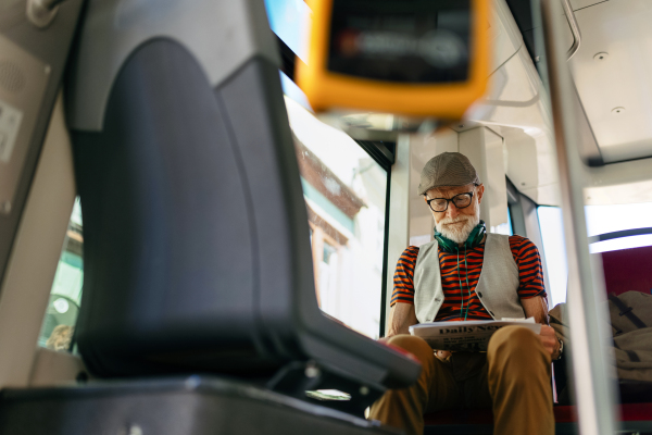 Elderly man traveling through the city by bus, reading a newspaper. Senior city commuter taking tram to grocery store, using public transportation.