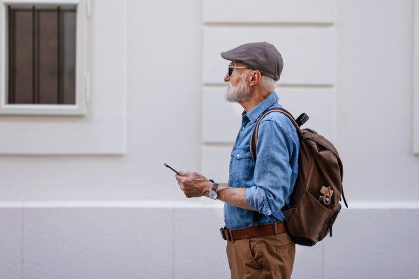 Side view of senior tourist exploring a new city, interesting places. Elderly man holding smartphone, looking for the route. Traveling and solo trips in retirement.