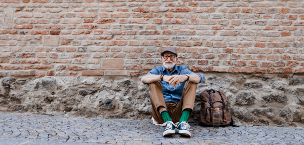 Banner of senior tourist exploring new city, interesting places. Elderly man with backpack sitting by brick wall, resting. Traveling and solo trips in retirement. Banner with copy space.