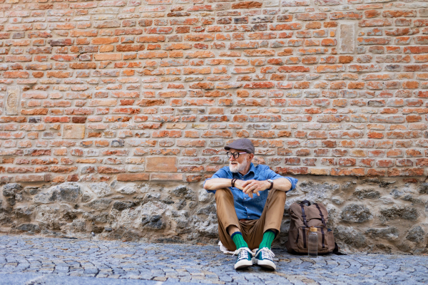 Portrait of senior tourist exploring new city, interesting places. Elderly man with backpack sitting by brick wall, resting. Traveling and solo trips in retirement, copy space.