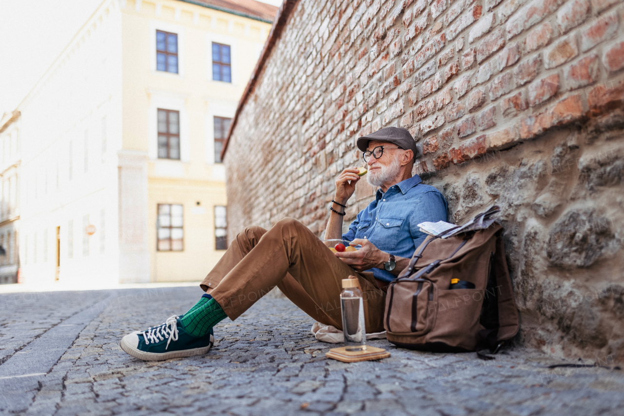 Elderly man with backpack sitting by the brick wall, reating healthy homemade snack and resting. Traveling and solo trips in retirement.