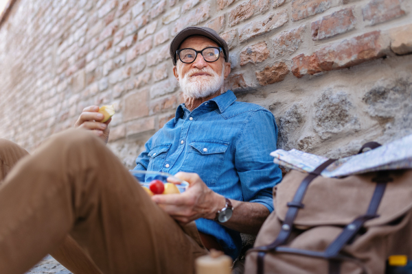 Elderly man with backpack sitting by the brick wall, reating healthy homemade snack and resting. Traveling and solo trips in retirement.