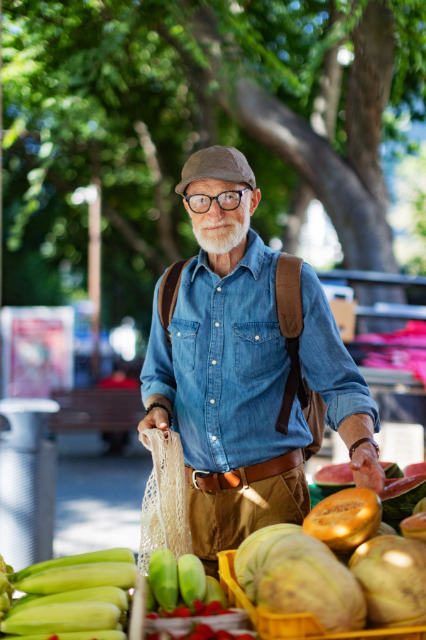 Portrait of senior man shopping at market in the city. Elderly man buying fresh vegetables and fruits from market stall.