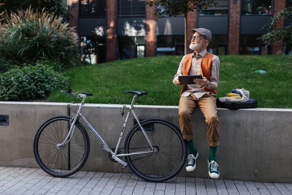 Stylish senior man sitting in the city park, holding tablet, shopping online. Concept of digital literacy, seniors using digital technologies.