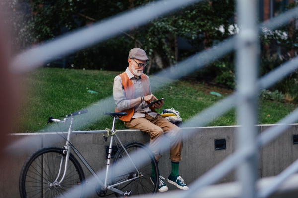 Stylish senior man sitting in the city park, holding tablet, shopping online. Concept of digital literacy, seniors using digital technologies.