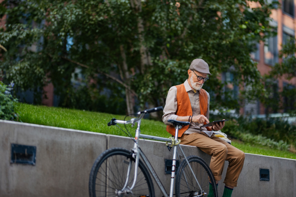 Stylish senior man sitting in the city park, holding tablet, shopping online. Concept of digital literacy, seniors using digital technologies.