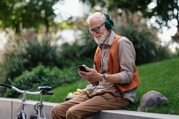 Stylish senior man sitting in the city park, listening to music via wireless headphones. Concept of old man young at heart.