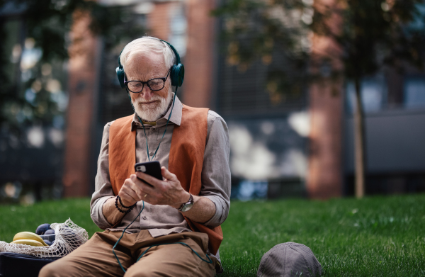 Stylish senior man sitting in the city park, listening to music via wireless headphones. Concept of old man young at heart.