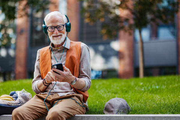 Portrait of stylish senior man sitting in the city park, listening to music via headphones. Concept of old man young at heart.