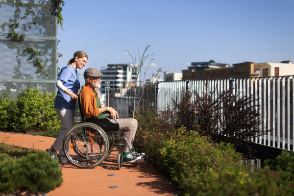 Nurse pushing senior man in a wheelchair. Female caregiver and elderly man enjoying a warm autumn day in nursing home, residential care home.