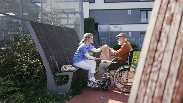 Nurse eating lunch or dinner outdoors with senior man in wheelchair. Female caregiver and elderly man enjoying a warm autumn day in nursing home, having picnic.